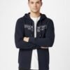 Men’s Navy Blue Zip-Up Hoodie with Logo Print Man wearing a navy blue zip-up hoodie with white printed logo, white t-shirt underneath, and black jeans, standing with arms crossed against a plain white background.
